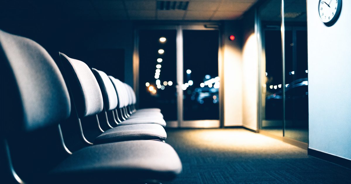 A quiet, clean emergency department waiting area at night with rows of gray chairs, a wall clock and glass doors with city lights outside.