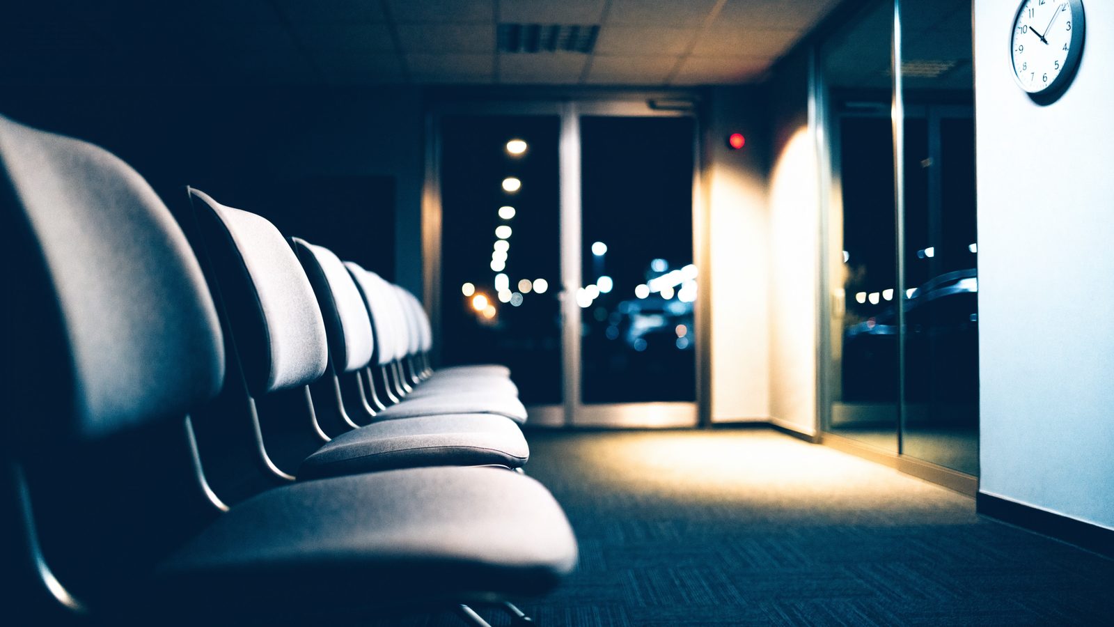 A quiet, clean emergency department waiting area at night with rows of gray chairs, a wall clock and glass doors with city lights outside.